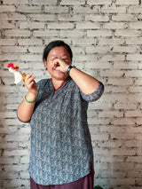 A handmade wool felt chicken finger puppet is held up by a smiling Nepalese female artisan, shown against a rustic white brick wall. This unique felt chicken puppet features a soft white body, a vibrant red crest, and yellow beak and legs. Each puppet is skillfully handcrafted, highlighting its unique artisanal quality.