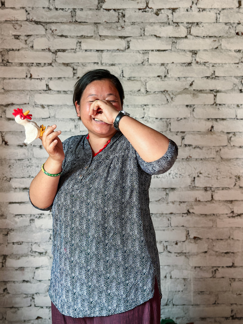 A handmade wool felt chicken finger puppet is held up by a smiling Nepalese female artisan, shown against a rustic white brick wall. This unique felt chicken puppet features a soft white body, a vibrant red crest, and yellow beak and legs. Each puppet is skillfully handcrafted, highlighting its unique artisanal quality.