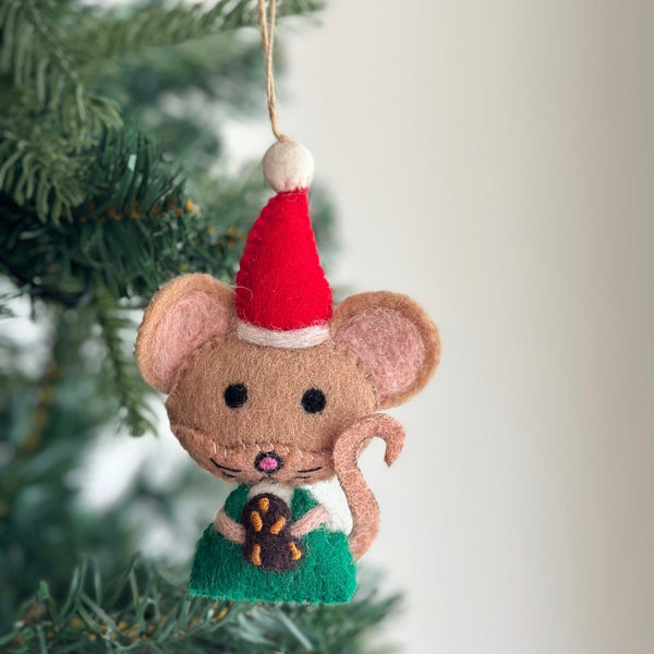 A handmade felt Christmas Cookie Mouse ornament hangs on a Christmas tree. The charming light brown mouse features round black eyes, a pink nose, and a stitched mouth. Dressed in a green felt outfit and a red Santa hat with a white pompom, it holds a chocolate-dipped pretzel cookie. A jute string on its hat allows for easy hanging.