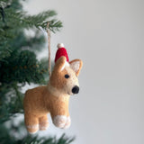 A needle felted Corgi Christmas ornament hangs gracefully from a Christmas tree branch, set against a soft, airy backdrop. The light brown Corgi features white paws and inner ears, and is adorned with a festive red Christmas hat. It is suspended by a rustic jute string.