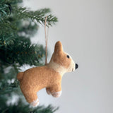 A wool felt Christmas Corgi ornament hangs gracefully on the green branches of a Christmas tree, captured in a close-up shot. This charming corgi ornament features a light brown body with a cream-colored chest and paws, emphasizing its adorable big butt. It is adorned with a festive Christmas hat and has a jute string attached for easy hanging, enhancing its rustic charm.