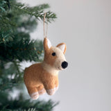 A handcrafted wool felt Corgi Christmas ornament hangs on a lush green Christmas tree. This light brown and creamy white needle felted corgi dog ornament features a shiny black nose, perky ears, white-tipped paws, and an adorably emphasized big butt. It is adorned with a festive red and white Christmas hat, bringing warmth and whimsy to holiday decor.