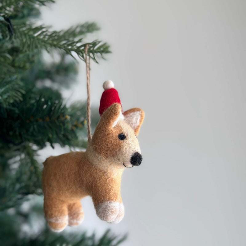 A charming felt corgi ornament, also a festive Christmas dog ornament, dangles from a green Christmas tree branch against a soft white backdrop. Crafted from soft, light brown felt, this corgi wears a red Christmas hat with a white pompom. It has small upright ears, a tiny black nose, and white markings on its paws, adding a heartwarming touch to holiday decor.