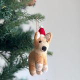 A needle felted corgi ornament, also known as a Christmas dog ornament, hangs festively on a Christmas tree. This charming corgi features a tan body with white paws and inner ears, black eyes and nose, and wears a red Christmas hat. It is suspended by a jute string, adding a joyful touch to the tree.