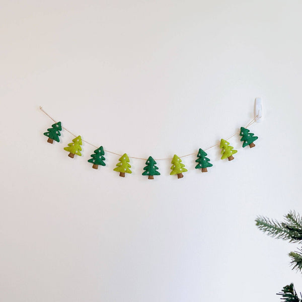 A felt Christmas Tree Garland with jute thread hangs gracefully on a clean white wall, with a hint of a real Christmas tree in the lower right corner. This festive garland features alternating green and light green felt Christmas tree shapes, each adorned with embroidered white snowflakes and brown tree trunks. The jute thread provides a rustic, natural texture.