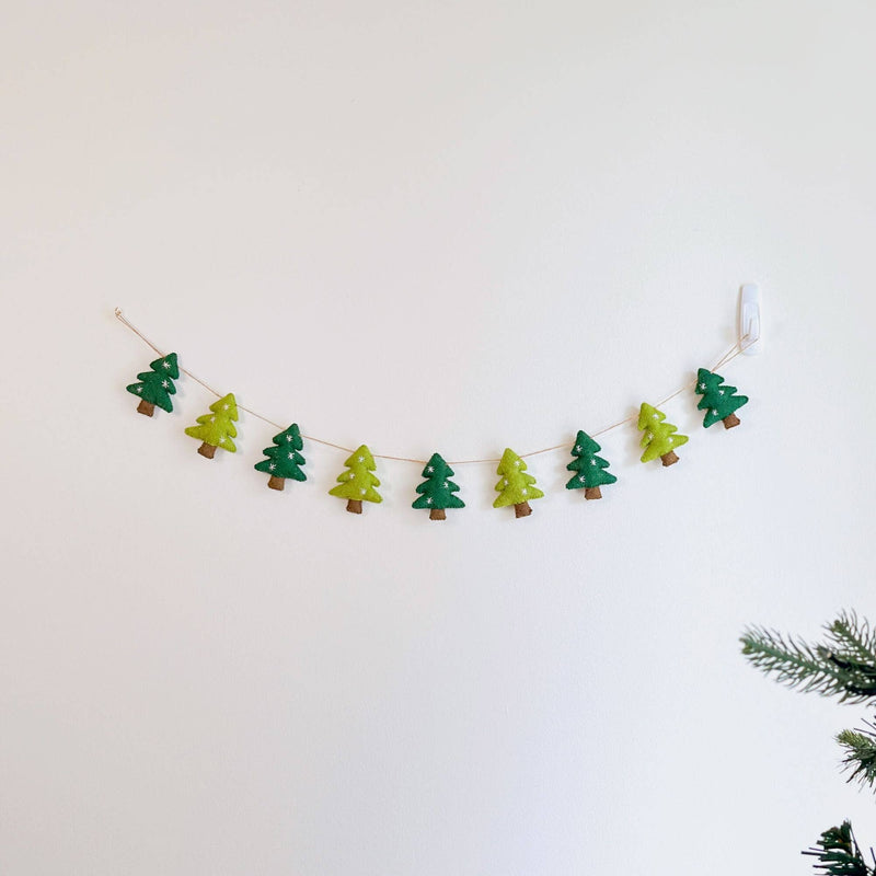 A felt Christmas Tree Garland with jute thread hangs gracefully on a clean white wall, with a hint of a real Christmas tree in the lower right corner. This festive garland features alternating green and light green felt Christmas tree shapes, each adorned with embroidered white snowflakes and brown tree trunks. The jute thread provides a rustic, natural texture.
