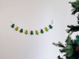A handmade Christmas tree felt garland, featuring green tree shapes, is strung across a plain white wall. Each felt tree has a small brown trunk and tiny white star details, connected by a natural jute thread, adding a rustic and festive touch.