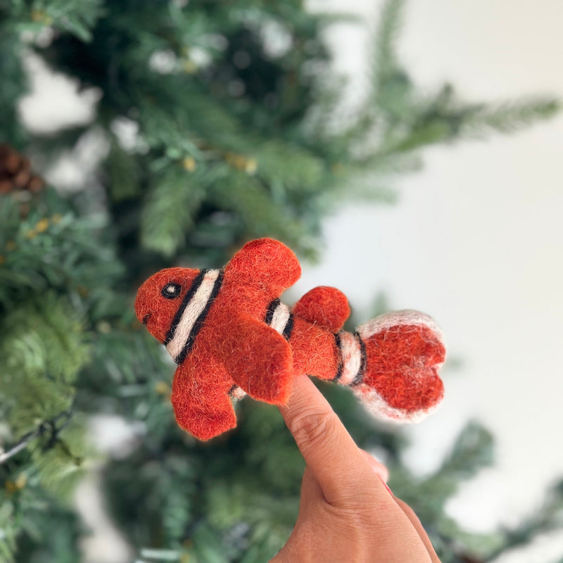 An orange and white wool felt clown fish finger puppet is perched on a finger, presented against a blurred green Christmas tree. This handmade finger puppet features distinctive black stripes and a whimsical googly eye, highlighting its textured, handcrafted quality.
