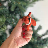 A handcrafted wool felt clown fish finger puppet is held by a hand, set against a soft-focus Christmas tree, evoking a cheerful, playful atmosphere. This unique felt finger puppet is bright orange with distinctive white and black stripes, featuring small fins, googly eyes, and a friendly smile.