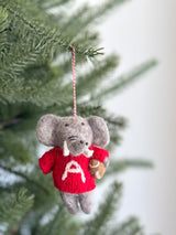 A handcrafted wool felt Alabama elephant ornament hangs from a blurred green Christmas tree. The gray elephant wears a bright red sweater featuring a prominent white letter 'A' and holds a football. It has black eyes and white tusks, suspended by a red and white cotton string.