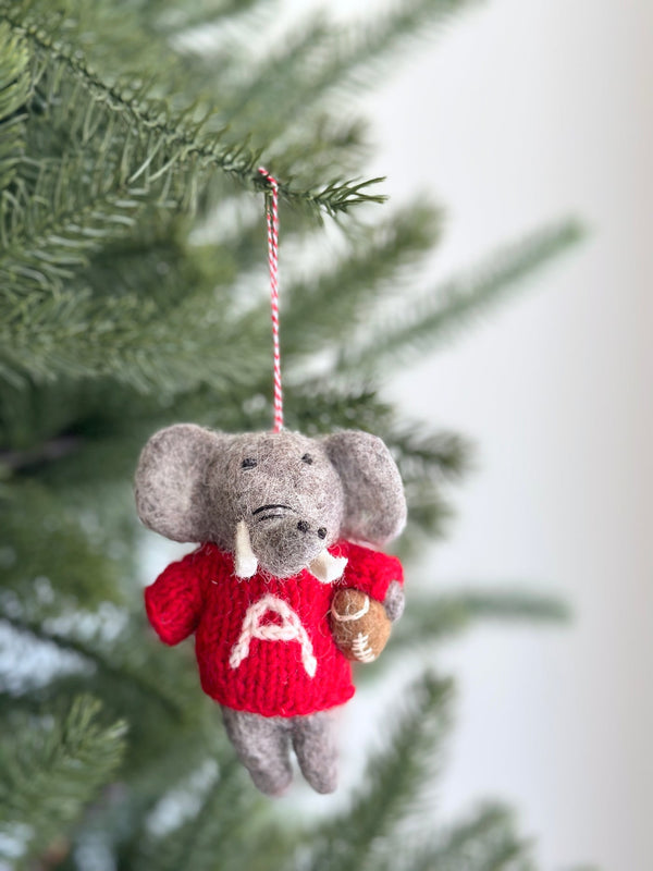 A handcrafted wool felt Alabama elephant ornament hangs from a blurred green Christmas tree. The gray elephant wears a bright red sweater featuring a prominent white letter 'A' and holds a football. It has black eyes and white tusks, suspended by a red and white cotton string.
