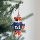 A spirited handcrafted wool felt Auburn Tiger ornament from the College Football Collection hangs on a Christmas tree. The felt Auburn Tiger wears a navy and burnt orange uniform with '02' on the front, holding a brown football. Its orange head features black stripes and white whiskers. A red and white string is attached to the top, ready for hanging.