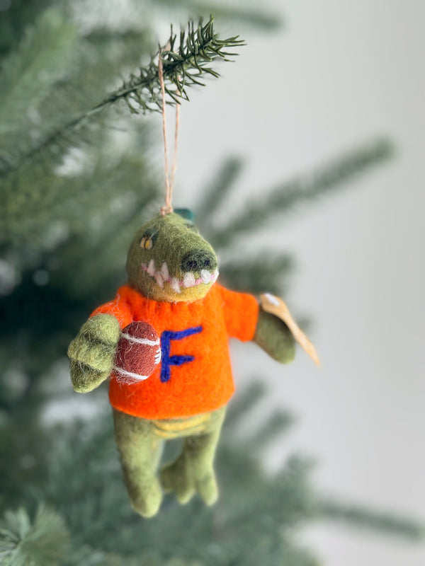 A green wool felt Florida gator Christmas tree ornament hangs on a green Christmas tree. The handcrafted felt gator wears a bright orange shirt with a blue 'F' and holds a brown football with white stripes. A jute string is attached for hanging, adding a festive touch to the holiday season.