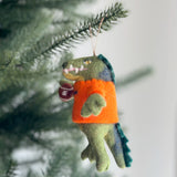 A handcrafted felt Florida Gator Christmas ornament hangs from a pine tree. The green gator felt ornament features a blue back and tail, an orange shirt, and holds a dark brown football, with a jute string attached for hanging.