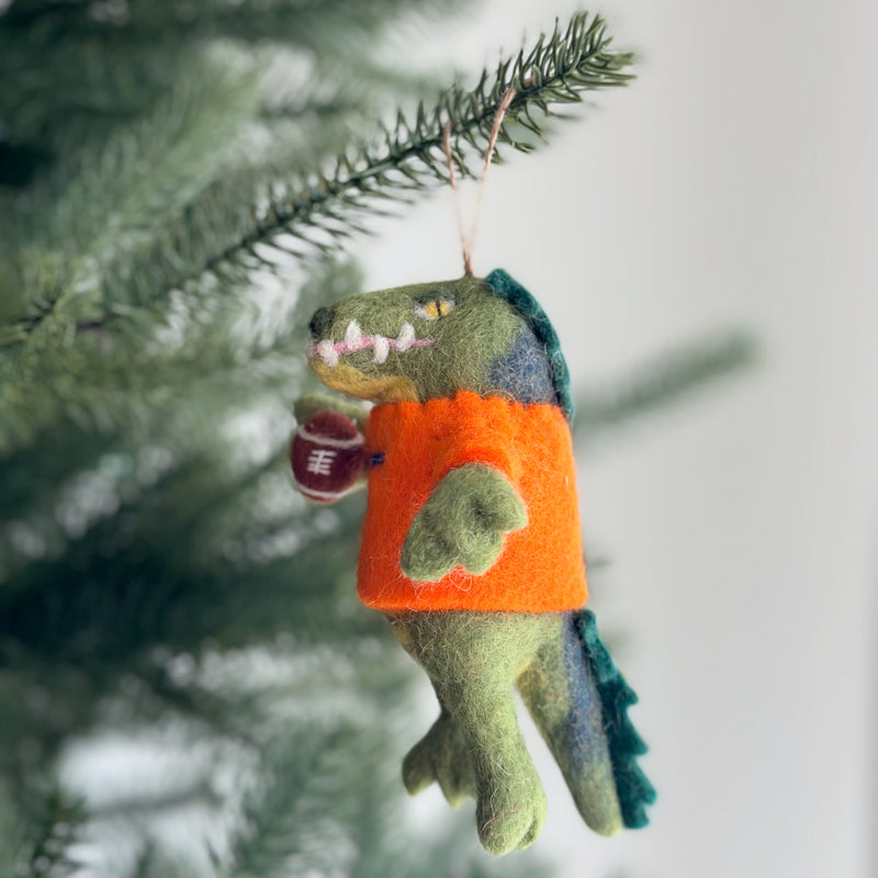 A handcrafted felt Florida Gator Christmas ornament hangs from a pine tree. The green gator felt ornament features a blue back and tail, an orange shirt, and holds a dark brown football, with a jute string attached for hanging.