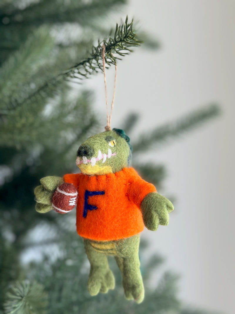 A handcrafted wool felt Florida Gator Christmas ornament, designed for team spirit, hangs on a festive Christmas tree. This playful green gator wears an orange sweater with a blue 'F' and holds a football, with yellow and pink details around its mouth. A jute string is attached for easy hanging.