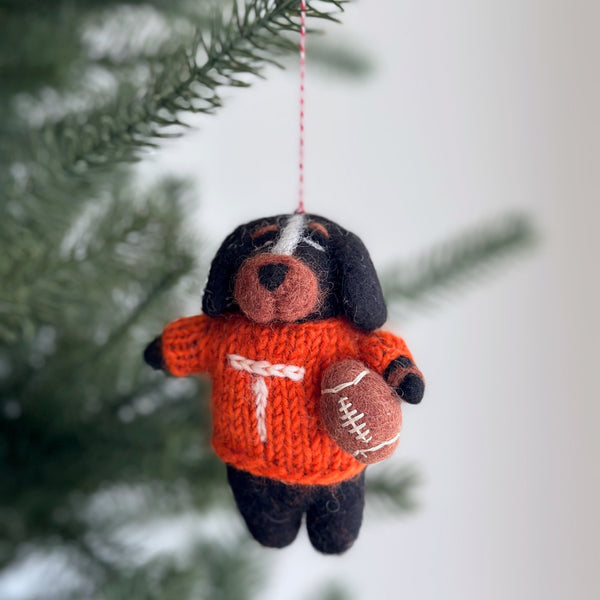 A handcrafted Tennessee Volunteers wool felt ornament hangs from a red and white cotton string against a soft, neutral background with hints of Christmas tree branches. This orange and white felt ornament showcases a bold 'T' design with visible hand-stitched details, perfectly capturing team spirit for any festive setting.