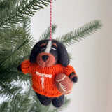 A Tennessee Volunteers wool felt dog ornament hangs from a pine tree branch, set against a bright, neutral backdrop. This handcrafted felt ornament depicts a black and brown dog wearing an orange sweater with a white 'T', holding a miniature football. It features hand-stitched details, floppy ears, and a light brown snout, exuding sportsmanship and holiday cheer. A red and white string is attached for display.