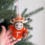 A handcrafted Texas Longhorns felt ornament is gently held by a hand, set against a blurred Christmas tree and a white wall, evoking a joyful, festive atmosphere. This unique wool felt ornament showcases the iconic burnt orange Longhorn silhouette, suspended by a red and white cotton string, embodying a bold and spirited essence.