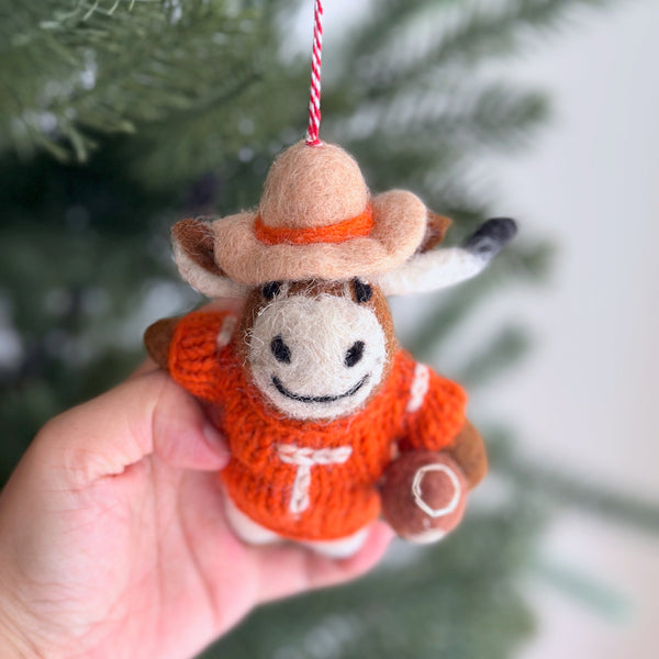 A handcrafted New Zealand wool felt Texas Longhorns ornament, an iconic piece from a college football collection, is held by a hand against a soft-focused background of green pine needles. The burnt orange ornament features the distinct Longhorns silhouette with a soft, textured surface and a spirited red and white cotton string for hanging.