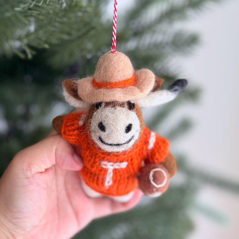 A handcrafted New Zealand wool felt Texas Longhorns ornament, an iconic piece from a college football collection, is held by a hand against a soft-focused background of green pine needles. The burnt orange ornament features the distinct Longhorns silhouette with a soft, textured surface and a spirited red and white cotton string for hanging.
