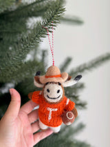 A Texas Longhorns felt ornament is held by a female hand, set against a soft-focus Christmas tree backdrop. This handcrafted, needle-felted wool felt ornament features the iconic Longhorn silhouette in burnt orange. The unique Christmas ornament hangs from a festive red and white cotton string.