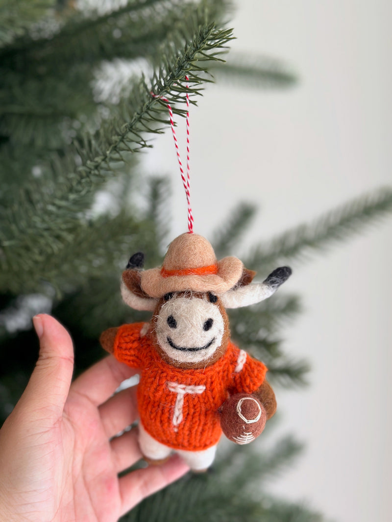 A Texas Longhorns felt ornament is held by a female hand, set against a soft-focus Christmas tree backdrop. This handcrafted, needle-felted wool felt ornament features the iconic Longhorn silhouette in burnt orange. The unique Christmas ornament hangs from a festive red and white cotton string.