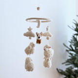 A wool felt Corgi and Sheep baby mobile is presented against a stark white background with a blurred green tree. This handmade nursery mobile features a playful brown and white corgi, cream sheep with pink ears and black stitched eyes and mouths, stars, clouds, and felt balls. All needle-felted from 100% wool, these charming elements hang from a circular wooden frame, with an extra felt ball suspended above.