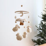 A Corgi and Sheep wool felt baby mobile hangs near a green Christmas tree against a bright, minimalist background, creating a whimsical and comforting atmosphere. The handmade mobile features a brown and white felt corgi positioned above fluffy white felt sheep, beige clouds, stars, and several round, white felt balls. All elements are suspended from a circular wooden frame, enhancing its rustic charm.