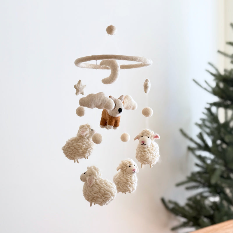 A Corgi and Sheep wool felt baby mobile hangs near a green Christmas tree against a bright, minimalist background, creating a whimsical and comforting atmosphere. The handmade mobile features a brown and white felt corgi positioned above fluffy white felt sheep, beige clouds, stars, and several round, white felt balls. All elements are suspended from a circular wooden frame, enhancing its rustic charm.