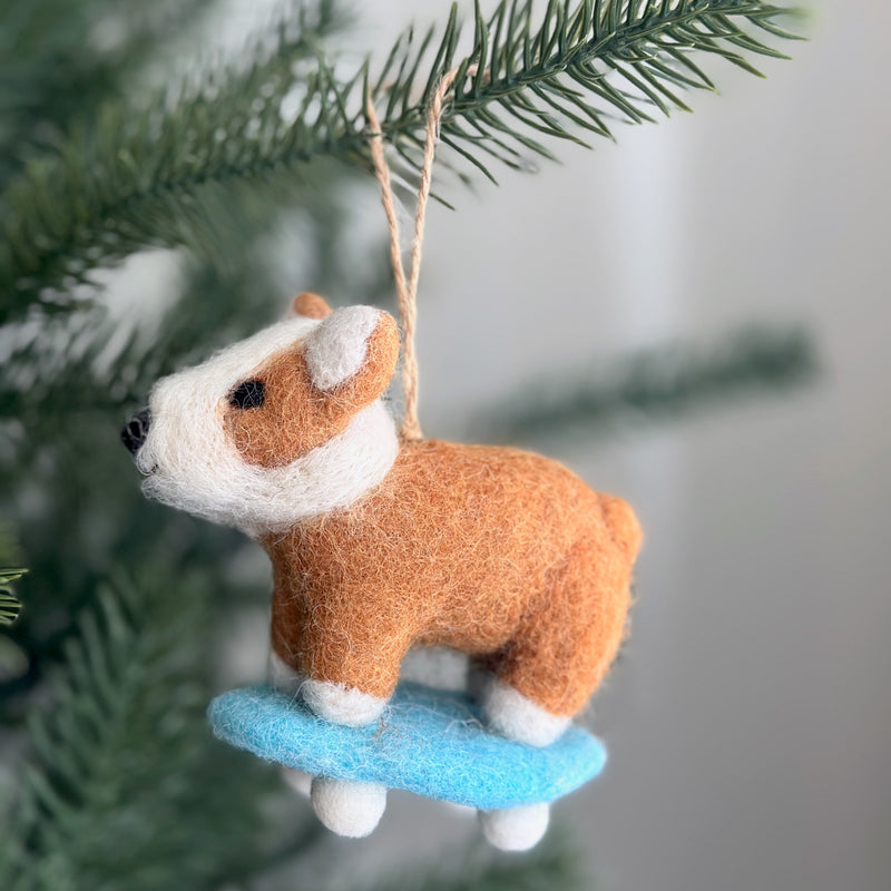 A handcrafted wool felt corgi skateboard ornament hangs playfully from a festive Christmas tree branch. The charming felt corgi features a light brown body with a white face, black nose, and eyes, perched on a light blue skateboard with white wheels. A jute string allows this unique Christmas ornament to be hung.