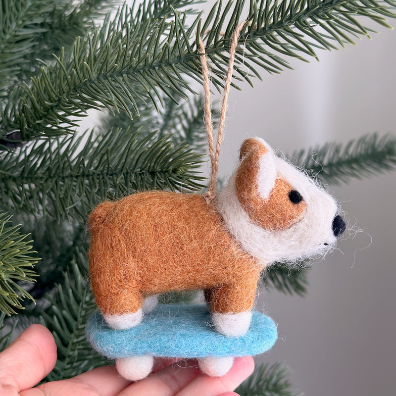 A handcrafted wool felt corgi on a skateboard ornament is held by a hand against a Christmas tree. This charming felt corgi features a brown body with white ears and a fluffy white chest, confidently riding a blue skateboard with white wheels. A jute string is attached for hanging this unique Christmas tree ornament.