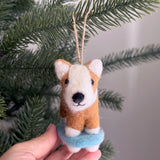 A felt Corgi on a skateboard ornament is gently held by a hand, poised against the soft green branches of a Christmas tree. This needle-felted wool felt Corgi features a white face and ears with a light brown body, riding a light blue skateboard. It is suspended by a jute string, adding playful charm to holiday decor.