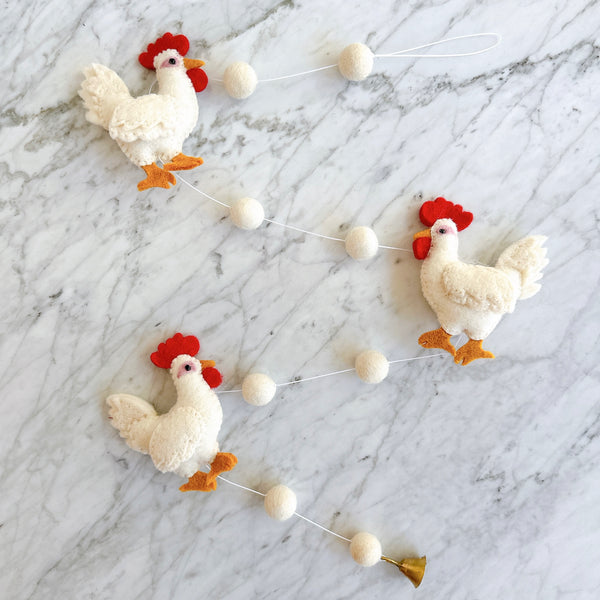 A cozy homestead wool felt chicken garland is displayed on a light marble background, evoking a calm, pastoral atmosphere. The garland features three white felt chickens, each with a red crest, orange beak, and orange feet. White felt balls are evenly spaced between the chickens, and a copper bell hangs from one end.