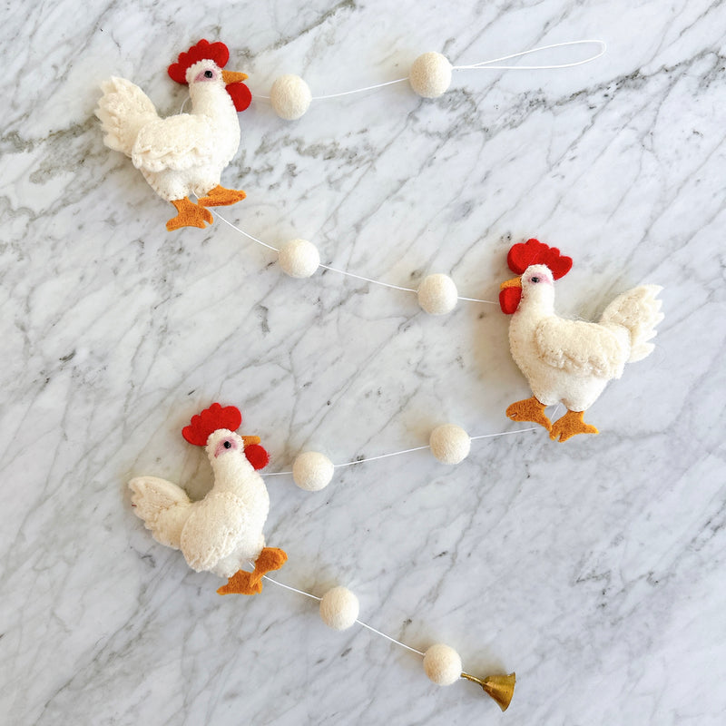 A cozy homestead wool felt chicken garland is displayed on a light marble background, evoking a calm, pastoral atmosphere. The garland features three white felt chickens, each with a red crest, orange beak, and orange feet. White felt balls are evenly spaced between the chickens, and a copper bell hangs from one end.