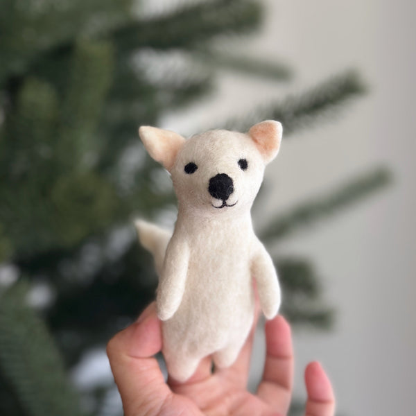 A handcrafted off-white needle-felted dire wolf finger puppet is held gently in a female hand, set against the softly blurred background of a festive Christmas tree. This unique felt puppet features small black eyes, a black nose, pointed ears with a pinkish inner hue, and a slender tail, designed perfectly for engaging finger play.
