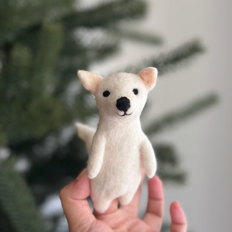 A handcrafted off-white needle-felted dire wolf finger puppet is held gently in a female hand, set against the softly blurred background of a festive Christmas tree. This unique felt puppet features small black eyes, a black nose, pointed ears with a pinkish inner hue, and a slender tail, designed perfectly for engaging finger play.