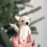 A handcrafted white wool felt dire wolf finger puppet is held in a female hand, with a blurred Christmas tree in the background. This unique dire wolf puppet features pointed ears with pink interiors, a black nose and eyes, and a slightly curved tail. Its soft, needle-felted texture evokes a cozy and imaginative feel for storytelling.