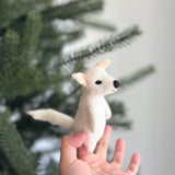 A light beige handmade wool felt dire wolf finger puppet is held upright in a female hand, appearing against a blurred background with a subtle Christmas tree. This unique felted finger puppet features a pointed tail, black eyes and nose, and peach inner ears, showcasing its soft wool felt texture.