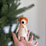 A creamy white handcrafted wool felt dog finger puppet with rust-colored ears and a golden bell collar is held in a hand against a soft-focused Christmas pine tree. This unique needle-felted dog puppet, handmade in Nepal, makes a festive storytelling toy.