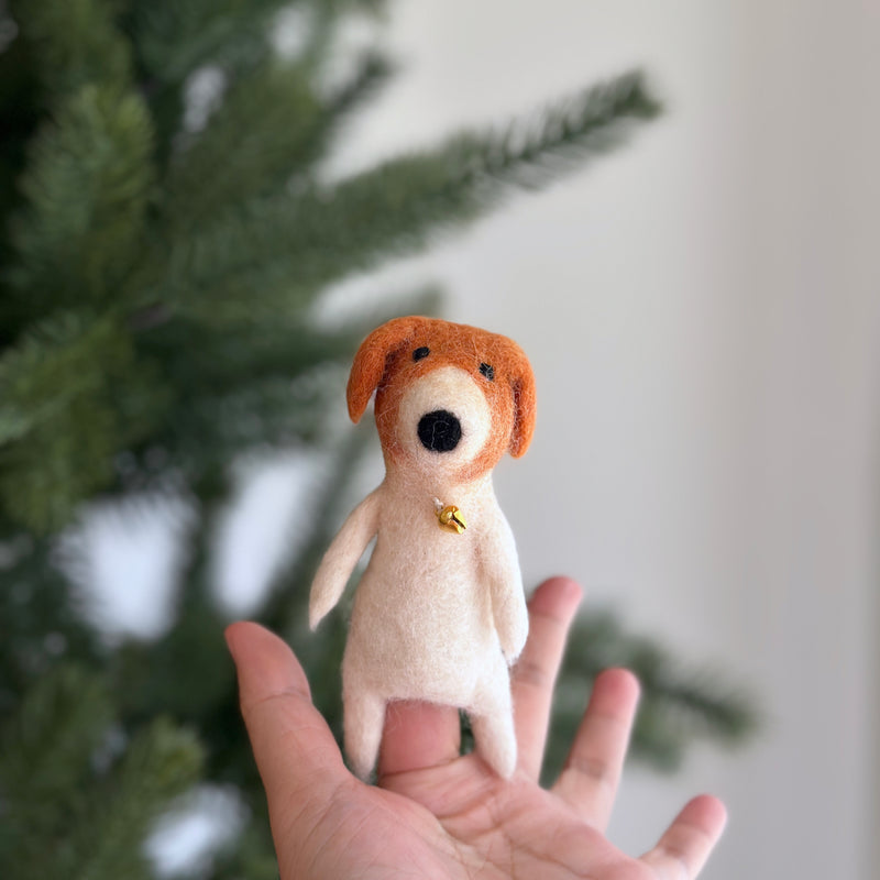 A creamy white handcrafted wool felt dog finger puppet with rust-colored ears and a golden bell collar is held in a hand against a soft-focused Christmas pine tree. This unique needle-felted dog puppet, handmade in Nepal, makes a festive storytelling toy.