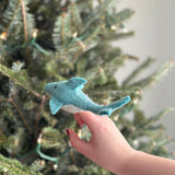 A blue and white wool felt dolphin finger puppet is held gently in a female hand, positioned in front of a soft-focused Christmas tree with subtle lights. This handcrafted fair-trade dolphin puppet features a light blue body, a white underbelly, small fins, and tiny black eyes. Its unique, needle-felted design makes it a charming toy for imaginative play.