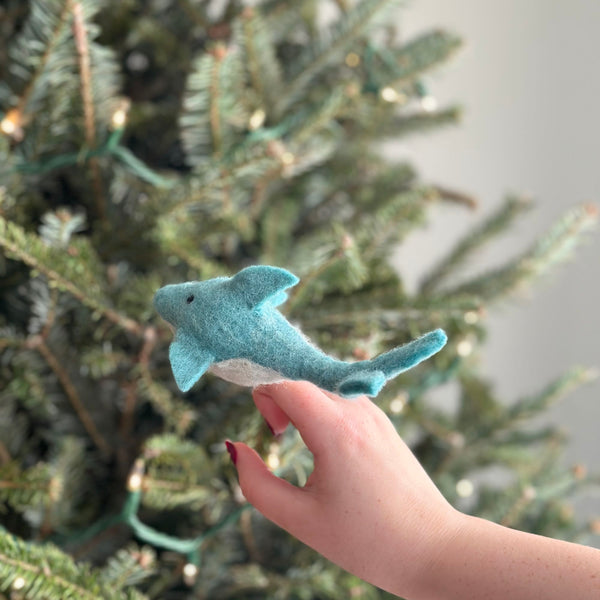 A blue and white wool felt dolphin finger puppet is held gently in a female hand, positioned in front of a soft-focused Christmas tree with subtle lights. This handcrafted fair-trade dolphin puppet features a light blue body, a white underbelly, small fins, and tiny black eyes. Its unique, needle-felted design makes it a charming toy for imaginative play.
