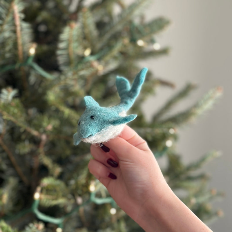 A festive handmade wool felt dolphin finger puppet is held by a hand with dark red nail polish, set against a blurred Christmas tree with white lights. This soft, dual-tone dolphin puppet has a light turquoise body, a white underbelly, fins, a tail, and expressive black eyes, showcasing its playful, handcrafted design.