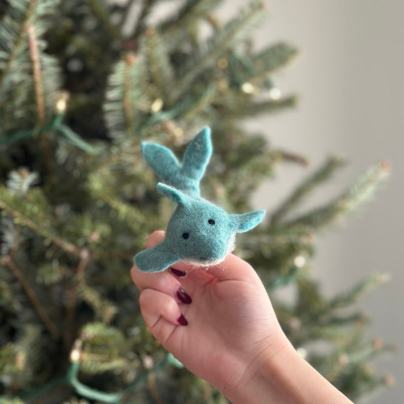 A light blue wool felt dolphin finger puppet is held in a female hand with dark red nail polish, against a blurred festive Christmas tree. This charming, handmade wool felt dolphin features two small black eyes, highlighting its unique design perfect for playful storytelling.