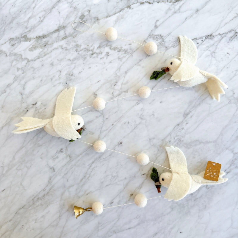 A serene felt Dove Garland is displayed on a white marble surface. This handcrafted felt garland features three white felt doves, each gently holding a small green olive branch in its beak. They are evenly spaced along a thin white string, with small round white felt balls in between. A small copper bell hangs at the bottom end, completing this unique design.