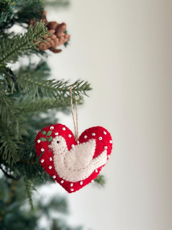 A handcrafted wool felt dove heart ornament hangs gracefully from a jute string on a Christmas tree branch, set against a softly blurred holiday background. The festive ornament features a red heart shape edged with small white bobbles, overlaid with a pure white stylized dove holding a sprig of green leaves in its beak.