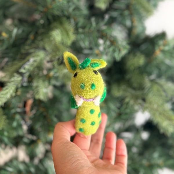 A whimsical green and yellow needle-felted dragon finger puppet is gently held in a woman's hand against a blurred Christmas tree, creating a festive mood. This handcrafted wool felt toy features small green wings, body spots, and tiny ears, perfect for holiday storytelling.