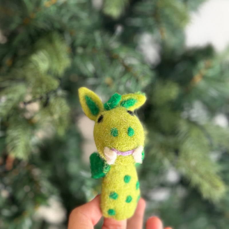 A cheerful, needle felted dragon finger puppet is perched against a green Christmas tree, creating a cozy, festive scene. This 100% wool dragon puppet is light green with darker green spots, accents, and white fangs. Its 3D design gives it a cute and friendly appearance, highlighting its playful design.