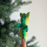 A handcrafted wool felt dragon finger puppet is worn on a finger, presented against a blurred green Christmas tree background. This vibrant, needle-felted dragon puppet has a light green body with darker green wings, tail, and a distinct spiky ridge running down its back. Its simple yet striking form makes it an engaging felt toy for kids.